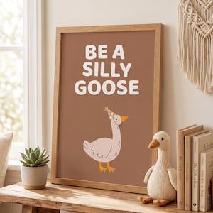 May include: A framed print with the words "BE A SILLY GOOSE" in white on a brown background. The print features a cartoon goose wearing a party hat. A knitted goose toy and books are on a wooden shelf.