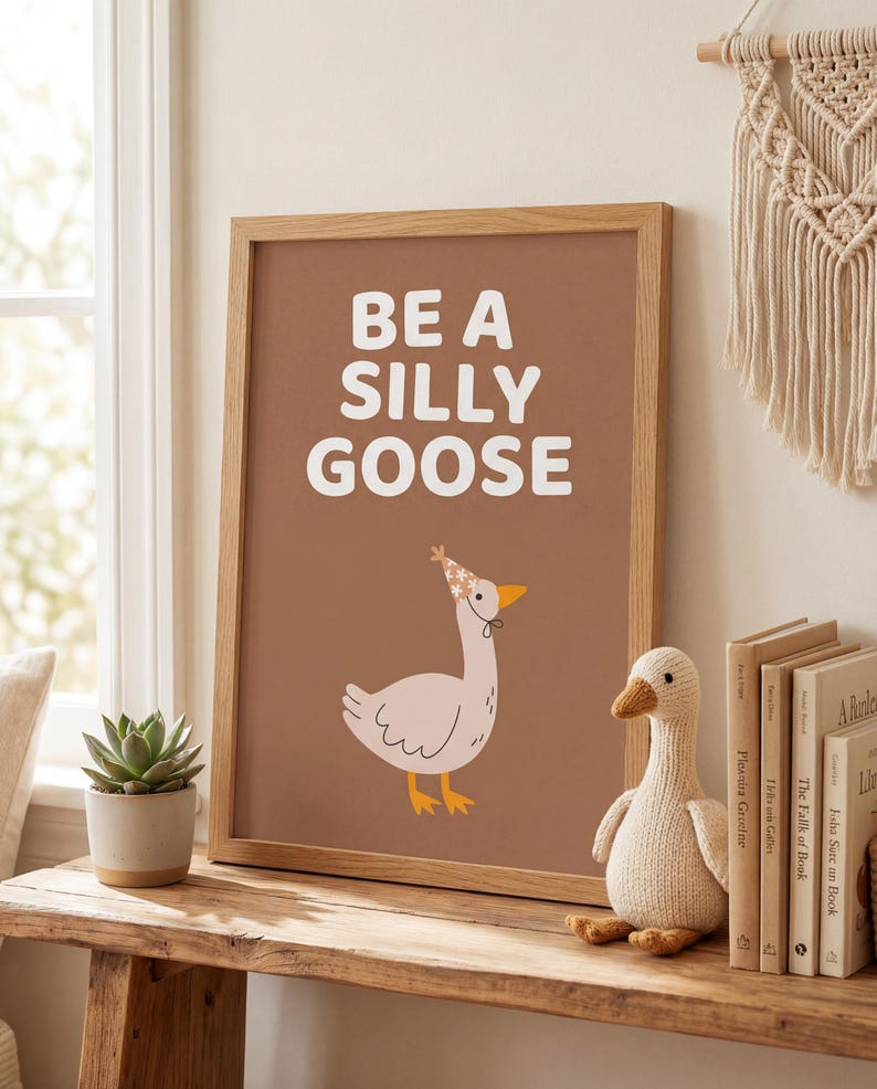 May include: A framed print with the words "BE A SILLY GOOSE" in white on a brown background. The print features a cartoon goose wearing a party hat. A knitted goose toy and books are on a wooden shelf.
