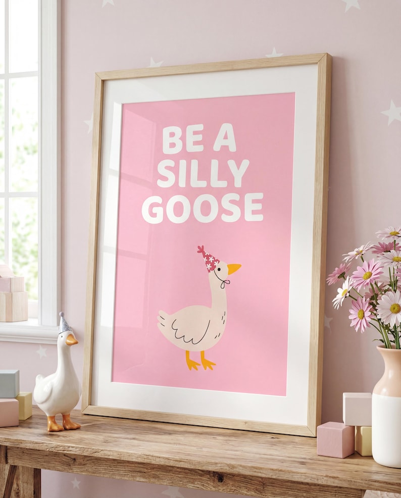 May include: A framed print with the words "BE A SILLY GOOSE" on a pink background. The print features a cartoon goose wearing a floral crown. The frame is light wood. A ceramic goose figurine and flowers are on a wooden surface.