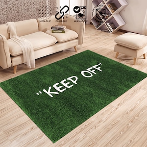 Op de afbeelding: Groen rechthoekig vloerkleed met de witte woorden "KEEP OFF". Het kleed heeft een grasachtige textuur en ligt op een houten vloer. Het kleed is antislip, duurzaam en wasbaar.