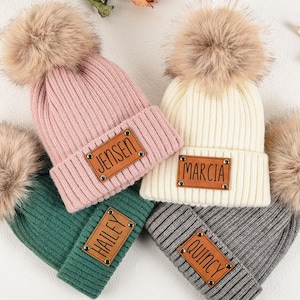 May include: Four knitted beanies in pink, cream, green, and gray, each with a faux fur pom-pom and a leatherette patch with a name. The names on the patches are "Jensen", "Marcia", "Hailey", and "Quincy".