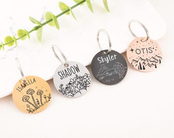 May include: Four round pet tags with silver key rings. The tags are in various colors: gold, silver, charcoal, and rose gold. Each tag has a different design, including mushrooms, flowers, a desert landscape, and mountains. The tags have names like "Isabella", "Shadow", "Skyler", and "Otis".