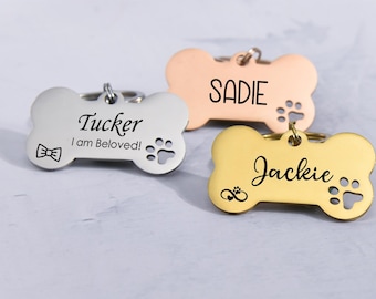 May include: Three personalised dog bone-shaped tags. The first tag is silver and says "Tucker I am Beloved!" with a bow tie and paw print. The second tag is rose gold and says "SADIE" with a paw print. The third tag is gold and says "Jackie" with an infinity symbol and a paw print.