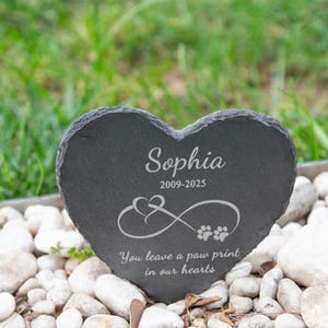 Puede incluir: Una l&aacute;pida de pizarra gris oscuro en forma de coraz&oacute;n con el nombre "Sophia" y las fechas "2009-2025" grabadas en blanco. Presenta un s&iacute;mbolo de infinito con huellas de patas y el texto "You leave a paw print in our hearts."
