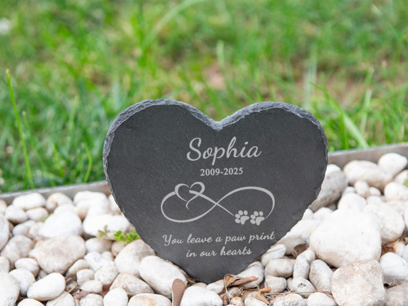 Puede incluir: Una l&aacute;pida de pizarra gris oscuro en forma de coraz&oacute;n con el nombre "Sophia" y las fechas "2009-2025" grabadas en blanco. Presenta un s&iacute;mbolo de infinito con huellas de patas y el texto "You leave a paw print in our hearts."