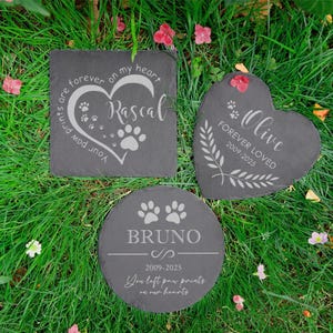 Puede incluir: Tres l&aacute;pidas conmemorativas para mascotas de pizarra gris oscuro en varias formas: cuadrada, coraz&oacute;n y c&iacute;rculo. Cada piedra est&aacute; grabada con el nombre de una mascota, huellas de patas y texto conmemorativo. Las piedras est&aacute;n sobre hierba verde con peque&ntilde;as flores rosas.