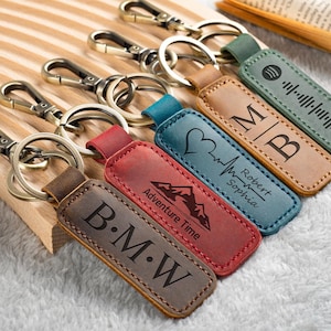 May include: A collection of personalized leather keychains with metal clasps. The keychains feature various designs, including initials, a mountain graphic, a heartbeat, and a music waveform. Colors include brown, red, blue, and green.