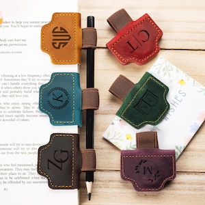 Op de afbeelding: Leren penhouders in verschillende kleuren, elk met een uniek monogram. De houders zijn bruin, blauwgroen, bordeauxrood, groen en paars. Een potlood wordt op zijn plaats gehouden door een elastiek. De houders liggen op een boek en een kaart.