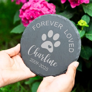 以下が含まれることがあります： 「FOREVER LOVED」の文字と、ハートの付いた足跡が刻まれた、丸い濃い灰色のスレートストーン。 「Charline」という名前と2009年から2025年までの年も刻まれています。 石は両手で持たれています。