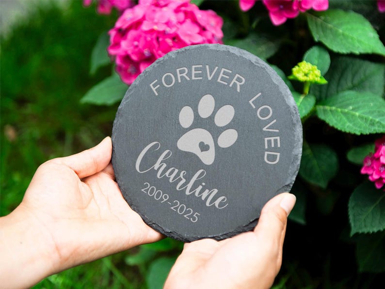 Puede incluir: Una piedra de pizarra redonda y gris oscuro con las palabras "FOREVER LOVED" y una huella de pata con un coraz&oacute;n. El nombre "Charline" y los a&ntilde;os 2009-2025 tambi&eacute;n est&aacute;n grabados. La piedra se sostiene con dos manos.