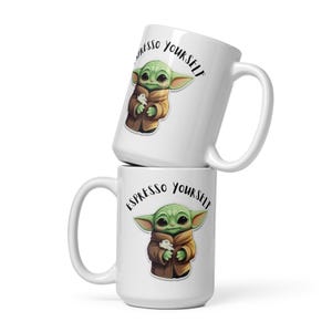 May include: White ceramic mug with a green character holding a small white object. The mug has the text "Espresso Yourself" in black lettering.