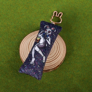 Puede incluir: Llavero rectangular con un personaje de dibujos animados con apariencia de conejo, vestido con un atuendo blanco y una falda a cuadros. El llavero tiene un anillo de metal dorado. El fondo es azul oscuro con un patrón de formas.