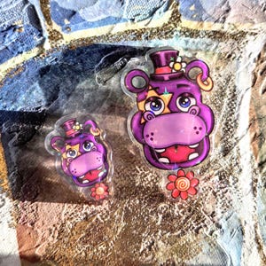 Puede incluir: Dos pegatinas transparentes con un personaje de hipopótamo morado con sombrero de copa, detalles de ojos en forma de estrella y una flor roja. Una pegatina es más grande que la otra. El fondo es una mezcla texturizada de azul, dorado y marrón.
