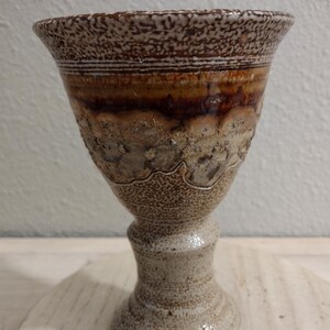 May include: A handcrafted ceramic goblet with a textured, speckled rim and a flared top. The goblet features a gradient of brown and tan hues, with a detailed, textured surface. The base is a pedestal design, adding to its elegant appearance.