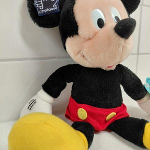 May include: A plush Mickey Mouse doll with black fur, a pink face, and large black ears. He wears red shorts with yellow buttons and yellow shoes. The doll has a tag that says "applause".