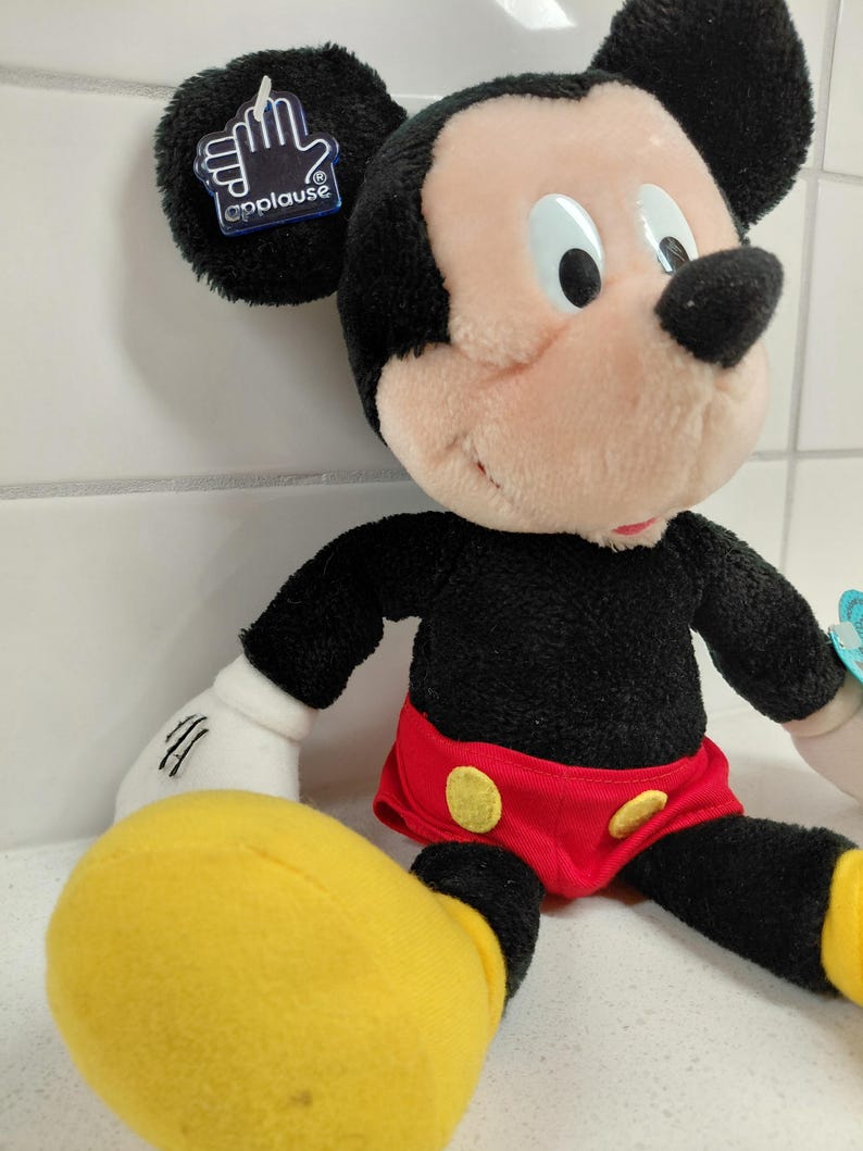 May include: A plush Mickey Mouse doll with black fur, a pink face, and large black ears. He wears red shorts with yellow buttons and yellow shoes. The doll has a tag that says "applause".