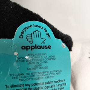 May include: A close-up of a Mickey Mouse plush toy tag. The tag is teal with white text and the word "applause" in red. The tag reads "Everyone loves to get" and includes cleaning instructions.