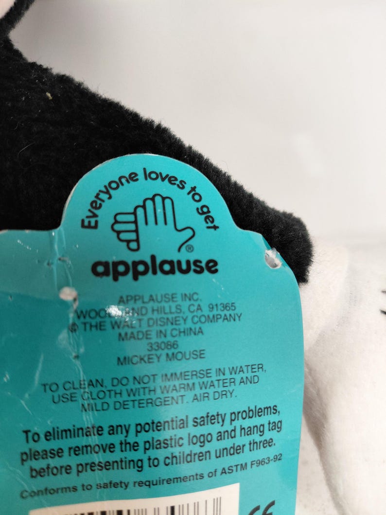 May include: A close-up of a Mickey Mouse plush toy tag. The tag is teal with white text and the word "applause" in red. The tag reads "Everyone loves to get" and includes cleaning instructions.