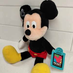 May include: A plush Mickey Mouse toy with black fur, large black ears, and a peach-colored face. He wears red shorts and yellow shoes. A tag reads "Mickey Unlimited" and features the Mickey Mouse logo.