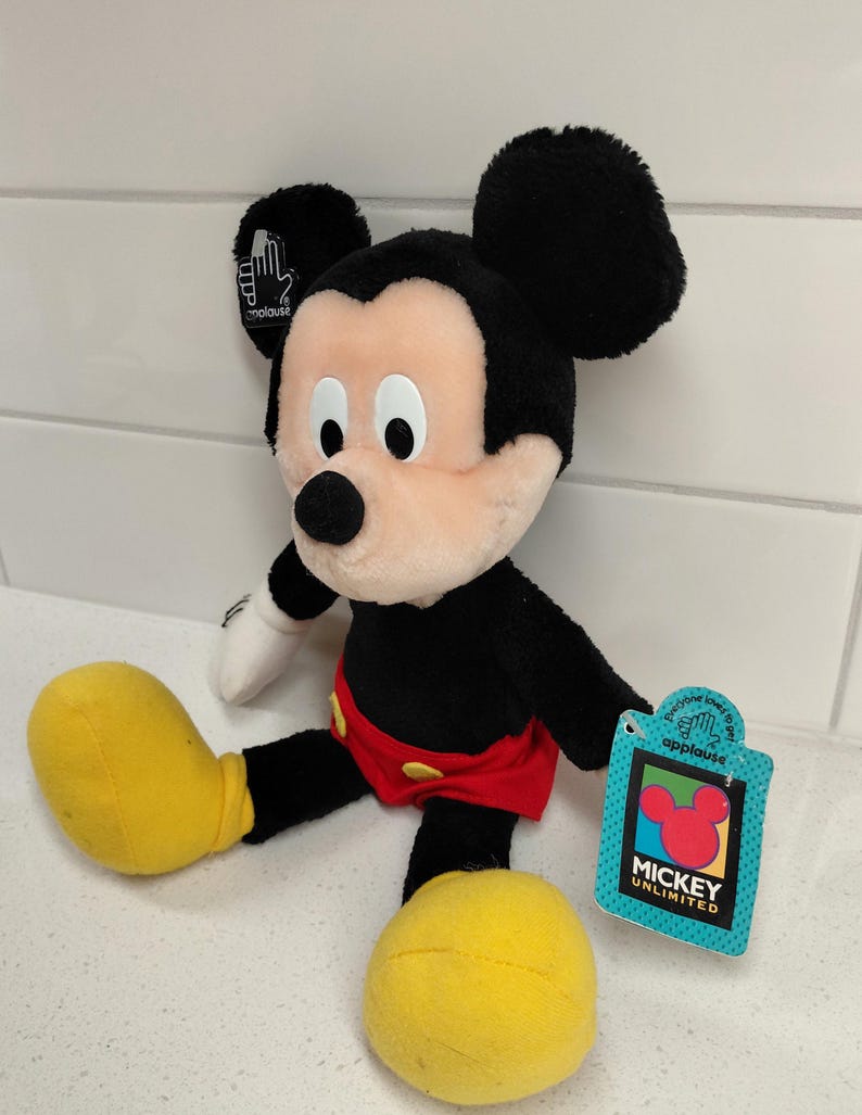 May include: A plush Mickey Mouse toy with black fur, large black ears, and a peach-colored face. He wears red shorts and yellow shoes. A tag reads "Mickey Unlimited" and features the Mickey Mouse logo.