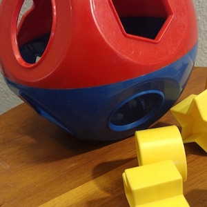 May include: A red, blue, and yellow shape sorter toy. The toy is a sphere with various shaped holes, including a circle, hexagon, and a rectangle. Yellow geometric shapes are scattered around the toy.