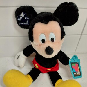 May include: A plush Mickey Mouse doll with black ears, a black body, and a peach-colored face. The doll has white gloves, red shorts, and yellow shoes. A blue tag with the Mickey Unlimited logo is attached to the doll.