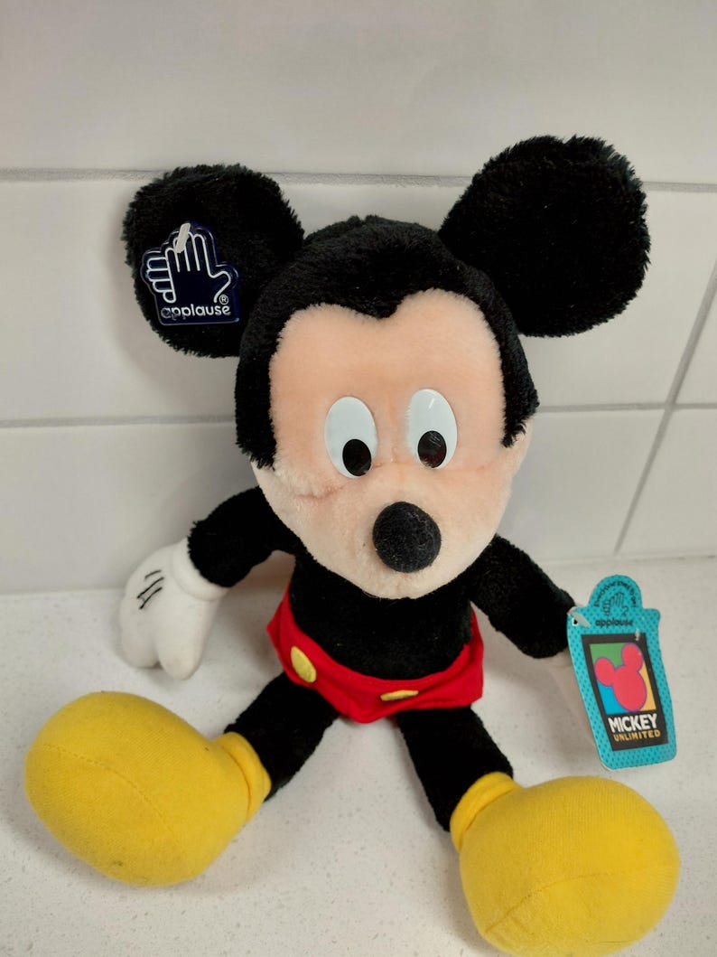 May include: A plush Mickey Mouse doll with black ears, a black body, and a peach-colored face. The doll has white gloves, red shorts, and yellow shoes. A blue tag with the Mickey Unlimited logo is attached to the doll.