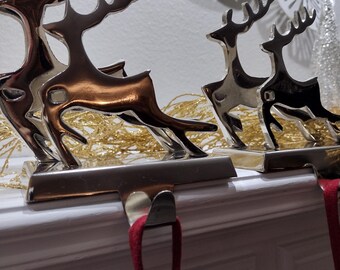 May include: Metal reindeer stocking holders in silver and copper tones. The reindeer are in a running pose, attached to a silver base with a hook for hanging stockings. Red stocking loops hang below. Decorative gold tinsel and white floral accents are in the background.