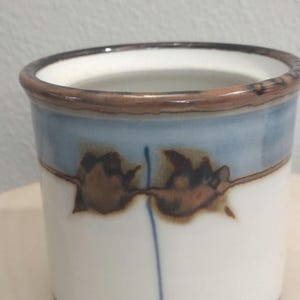 May include: A white ceramic mug with a brown rim and a blue and brown design. The design features two brown abstract shapes with a blue line in the center.