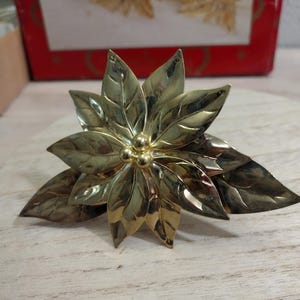 May include: A gold-colored poinsettia flower decoration with detailed leaves and a central cluster of small spheres. The flower is made of metal and sits on a light-colored wooden surface. A red and white box with gold flowers is in the background.