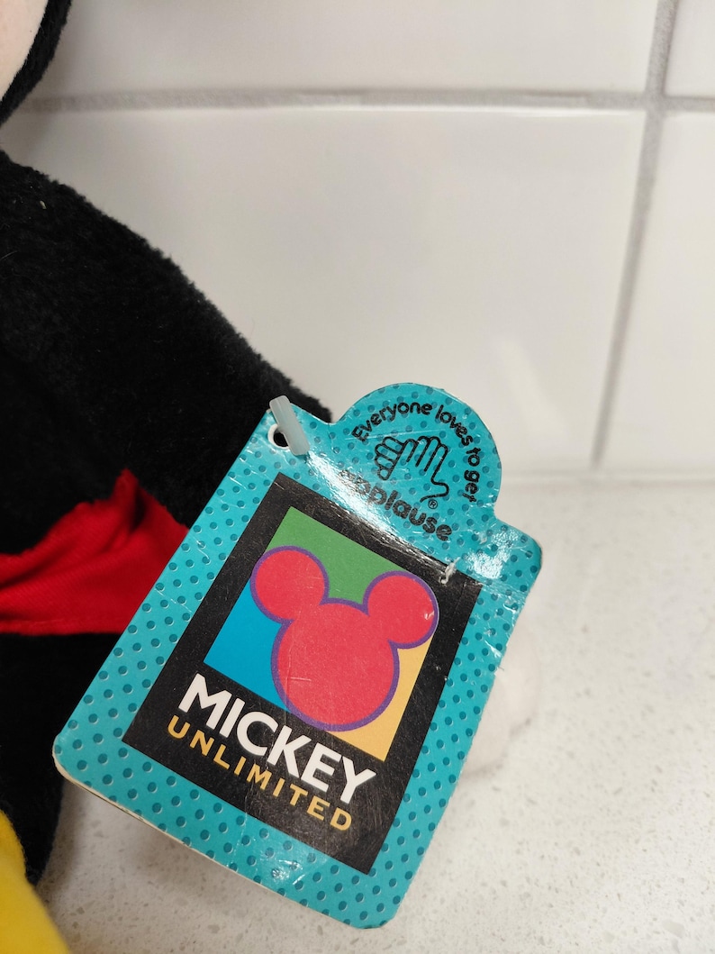 May include: A close-up of a Mickey Unlimited tag with a red Mickey Mouse head logo. The tag is teal with white polka dots and the text "Mickey Unlimited" in black. The tag also has the text "Everyone loves to get applause."