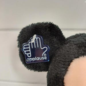 May include: Close-up of a black plush toy ear with a clear plastic tag. The tag features a hand outline and the word "applause". The toy has a soft, fuzzy texture and a light pink face.