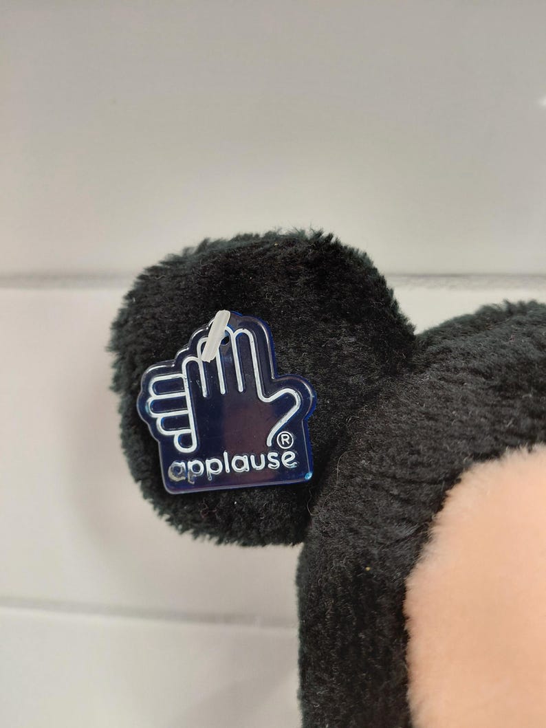 May include: Close-up of a black plush toy ear with a clear plastic tag. The tag features a hand outline and the word "applause". The toy has a soft, fuzzy texture and a light pink face.