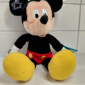 May include: A plush Mickey Mouse doll, featuring black ears, a black body, and a pink face. Mickey wears red shorts, yellow shoes, and white gloves. The doll has a tag that says "applause".