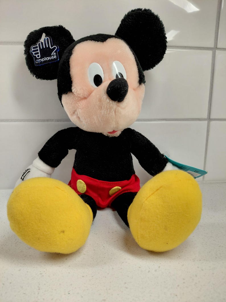 May include: A plush Mickey Mouse doll, featuring black ears, a black body, and a pink face. Mickey wears red shorts, yellow shoes, and white gloves. The doll has a tag that says "applause".
