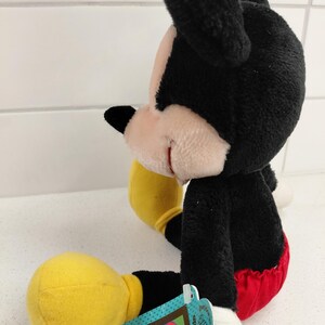 May include: A plush Mickey Mouse toy, featuring black fur, a pink face, and a red and white outfit. The toy has yellow gloves and a tag that reads "Mickey Unlimited".