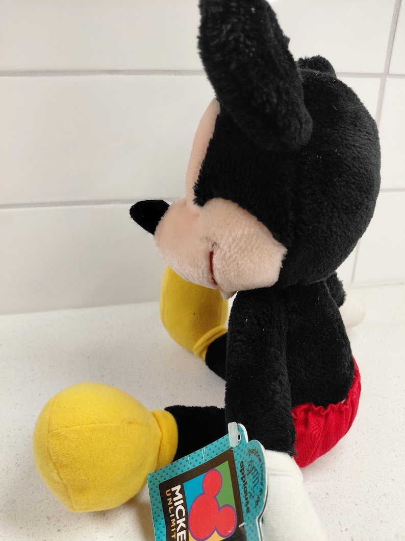 May include: A plush Mickey Mouse toy, featuring black fur, a pink face, and a red and white outfit. The toy has yellow gloves and a tag that reads "Mickey Unlimited".