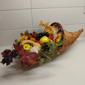 May include: A woven cornucopia overflowing with an assortment of faux fruits and foliage. The arrangement includes green and yellow apples, grapes, and fall leaves. The cornucopia is made of woven straw.