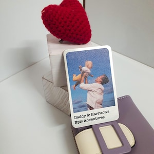 May include: A red crocheted heart sits on a small white block, next to a card with a photo of a man and a baby. The card reads "Daddy & Harrison's Epic Adventures." A purple wallet with a white strap is in the foreground.