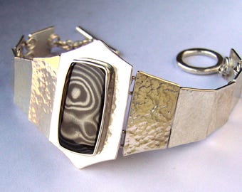 May include: A silver bracelet with a geometric design. The bracelet features a rectangular, black and white stone set in a silver frame. The bracelet is made of multiple silver links, some of which are textured.