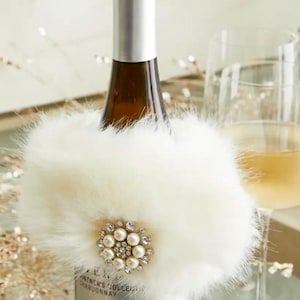 May include: A white wine bottle with a silver cap is adorned with a fluffy, cream-colored fur collar. The collar features a decorative brooch with pearls and crystals. A glass of white wine is in the background.