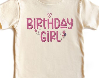 May include: A cream colored short-sleeved t-shirt with the text "Birthday Girl" in pink lettering. The text is decorated with pink hearts, stars, and balloons.