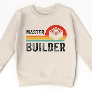 May include: Cream-colored sweatshirt with the words "MASTER BUILDER" in black, with a retro design of a red circle and a white Lego block. The design includes horizontal stripes in orange, yellow, and teal.