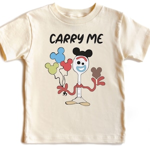 May include: A cream colored t-shirt with short sleeves features a cartoon character holding balloons. The character is a white spoon with a red and blue face, wearing a red and white shirt and black Mickey Mouse ears. The text "CARRY ME" is printed above the character.