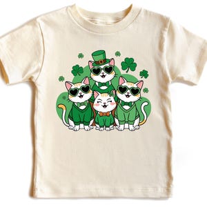 May include: A white t-shirt with a graphic of four cats wearing green shamrock hats and sunglasses. The cats are surrounded by green shamrocks.
