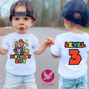 May include: Two white t-shirts with a Mario Brothers theme. One shirt features a graphic of Mario, Luigi, Toad, and Yoshi with the text "Birthday Boy". The other shirt has the text "Level 3" in colorful block letters.