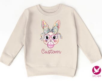 May include: A light beige sweatshirt with a colorful cartoon bunny wearing heart-shaped sunglasses and blowing a bubble. The text "Custom" is printed below the bunny.