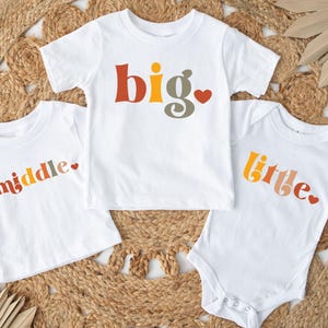 May include: Three white t-shirts with colorful text that reads "big", "middle", and "little" with a small red heart after each word. The shirts are on a woven straw background.