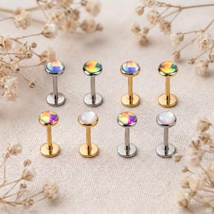 May include: Eight flat-back labret studs arranged in two rows. The studs feature circular tops with iridescent, multi-colored cabochon stones. Four studs have a silver-toned metal post and base, while the other four have a gold-toned metal finish.