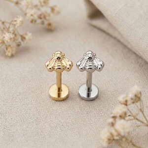 May include: Two flat back labret studs featuring a bee-shaped top. One stud is gold-toned and the other is silver-toned. Each piece consists of a circular base, a straight post, and a detailed bee ornament with textured wings and a rounded head.